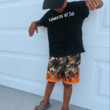 Child wearing a black cap, black t-shirt with text, and camouflage shorts with orange accents against a white garage door.