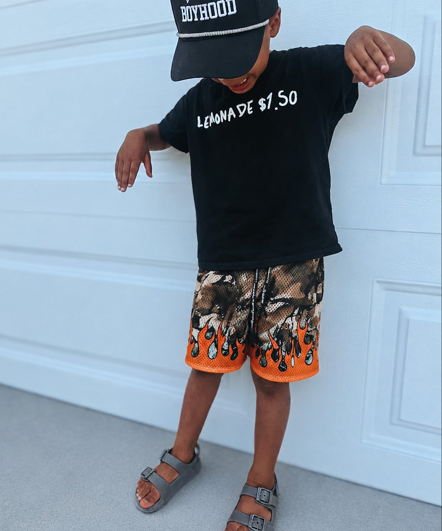 Child wearing a black cap, black t-shirt with text, and camouflage shorts with orange accents against a white garage door.