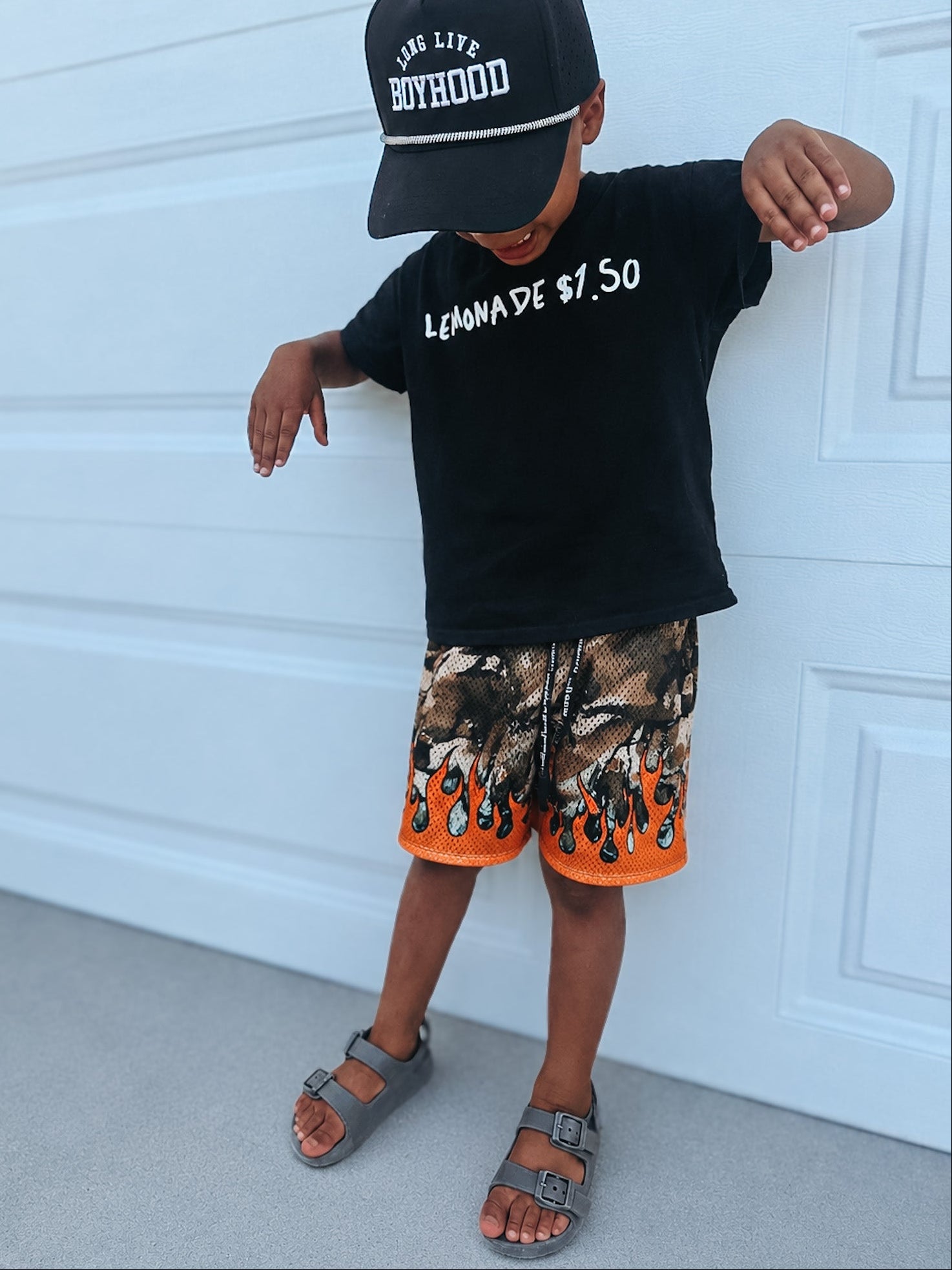 Child wearing a black cap, black t-shirt with text, and camouflage shorts with orange accents against a white garage door.