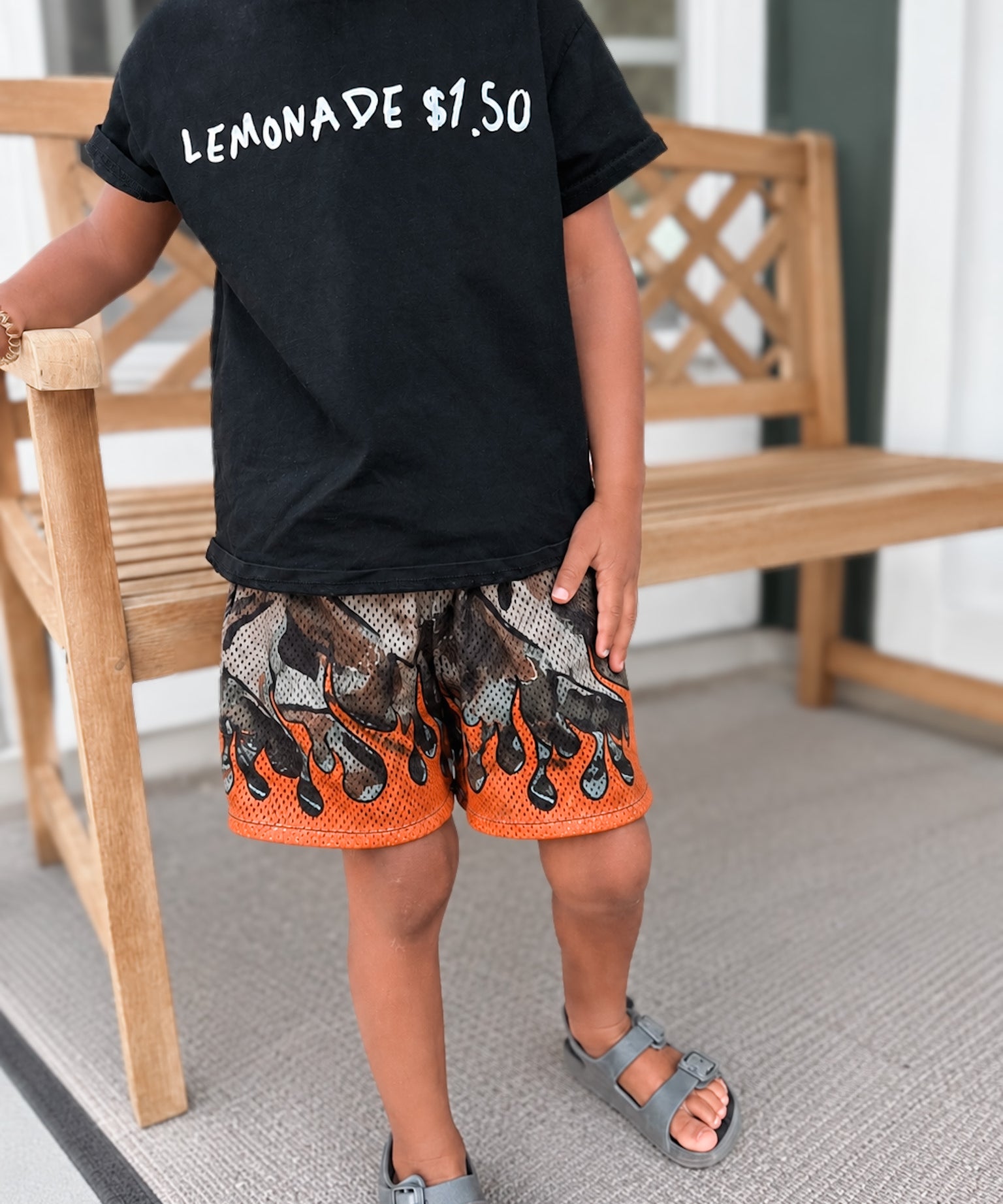 Child wearing a black t-shirt with 'LEMONADE $1.00' text, patterned shorts, and sandals on a porch.