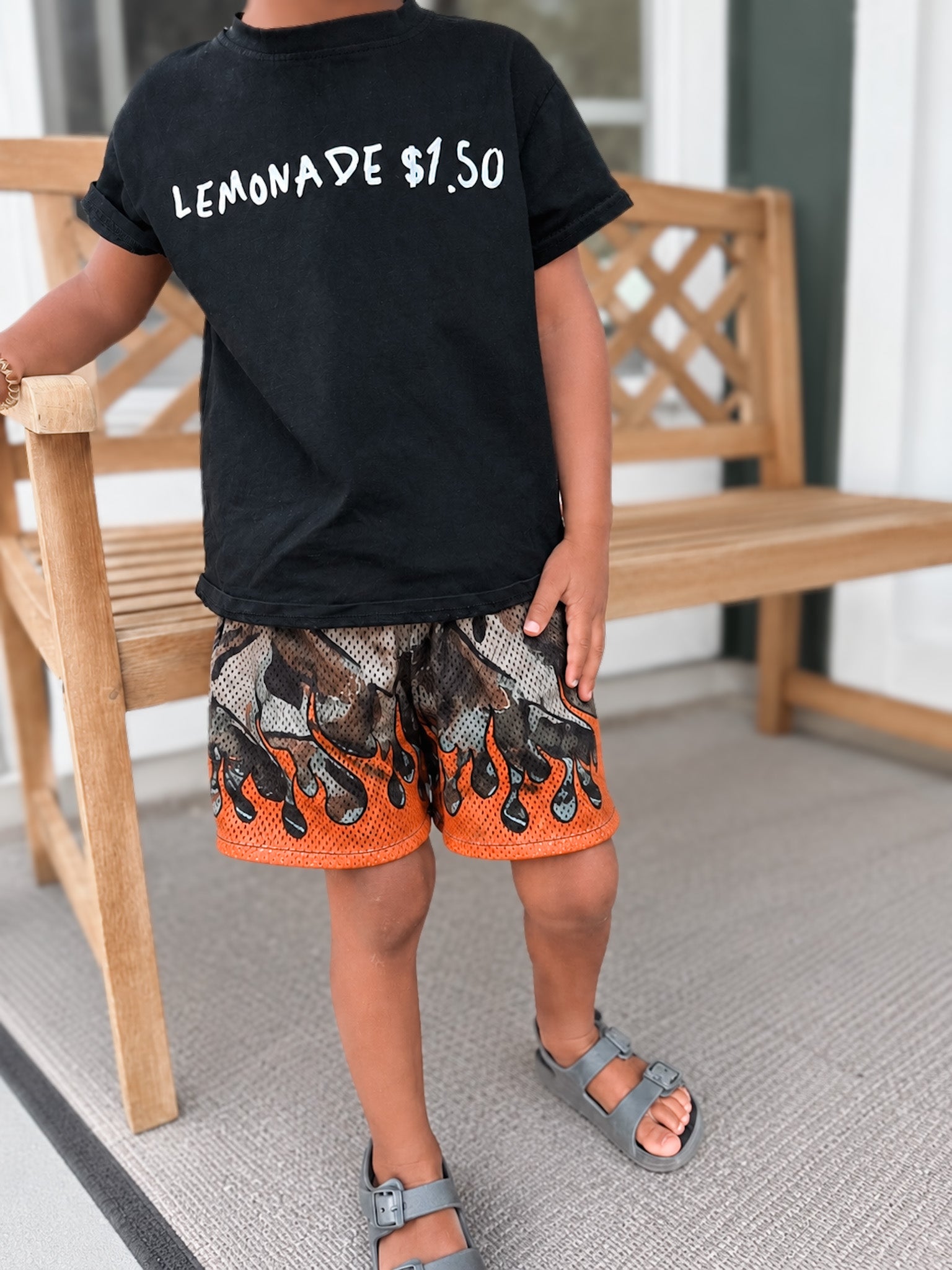 Child wearing a black t-shirt with 'LEMONADE $1.00' text, patterned shorts, and sandals on a porch.