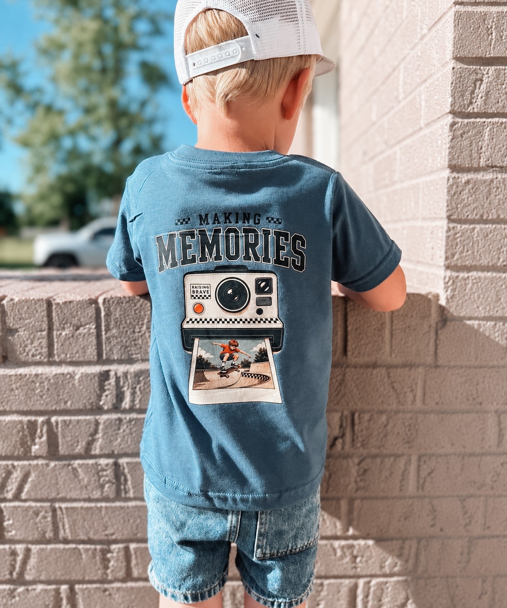 Child wearing a blue t-shirt with 'Making Memories' graphic leaning against a brick wall.