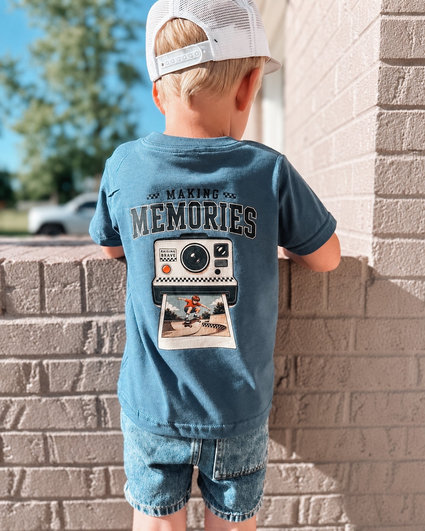 Child wearing a blue t-shirt with 'Making Memories' graphic leaning against a brick wall.
