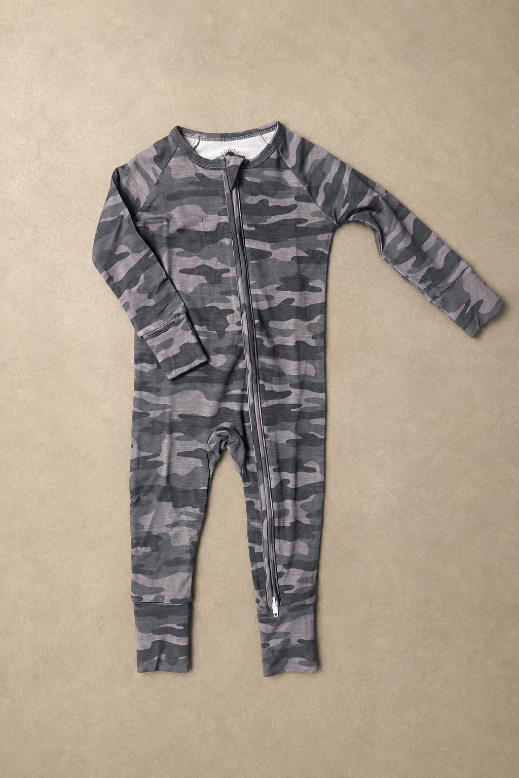 A long-sleeve baby footless one-piece jumpsuit with a gray and black camouflage pattern and a front zipper, laid flat on a textured beige background.