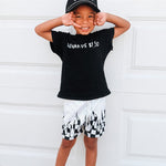Child wearing a black t-shirt with text and white shorts with black patterns, standing against a white wall.
