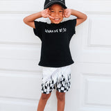 Child wearing a black t-shirt with text and white shorts with black patterns, standing against a white wall.