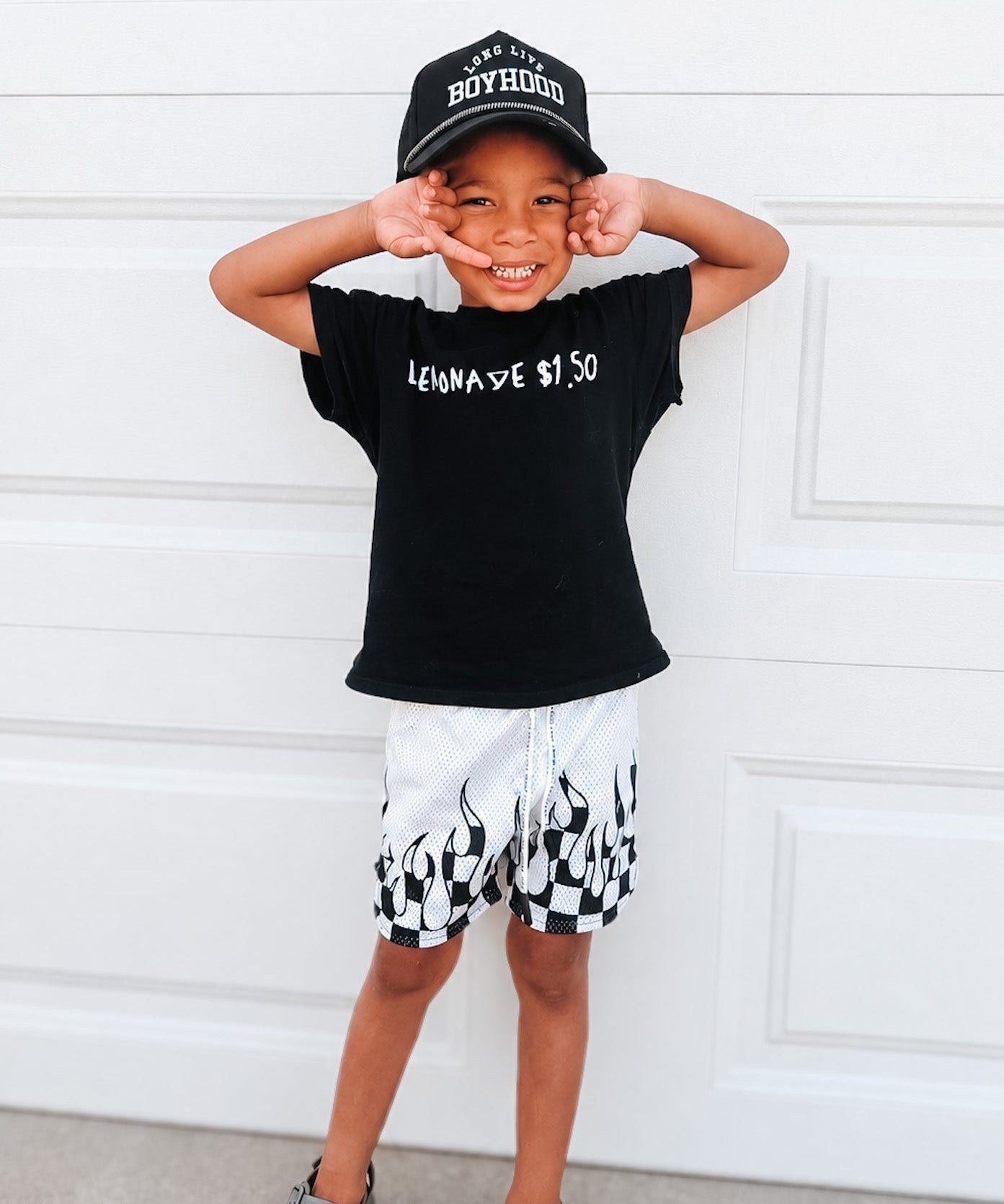 Child wearing a black t-shirt with text and white shorts with black patterns, standing against a white wall.