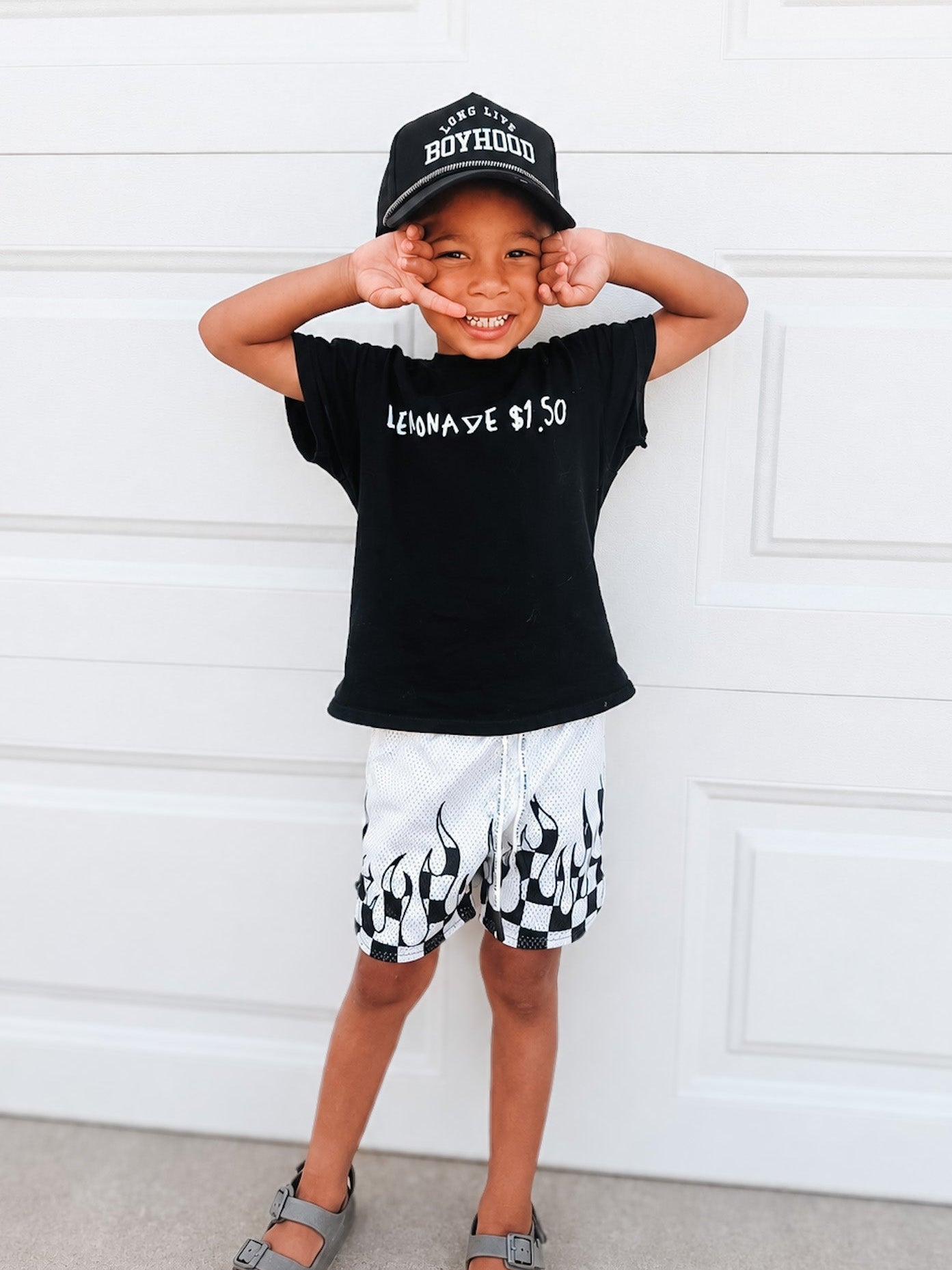 Child wearing a black t-shirt with text and white shorts with black patterns, standing against a white wall.