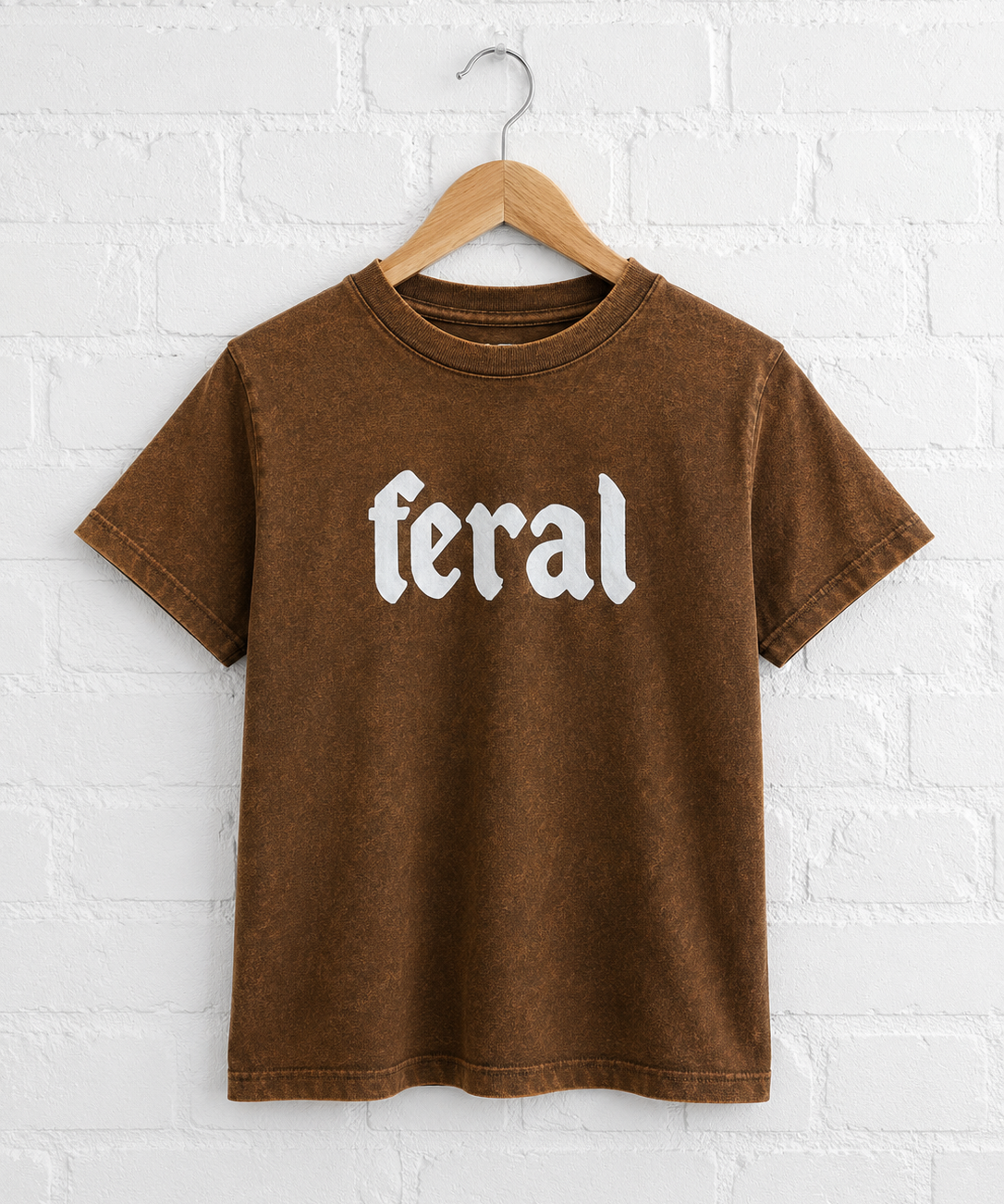 Brown t-shirt with 'feral' text on a hanger against a white brick wall.