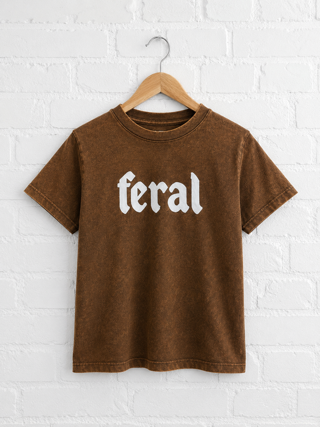 Brown t-shirt with 'feral' text on a hanger against a white brick wall.