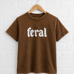 Brown t-shirt with 'feral' text on a hanger against a white brick wall.