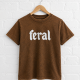 Brown t-shirt with 'feral' text on a hanger against a white brick wall.