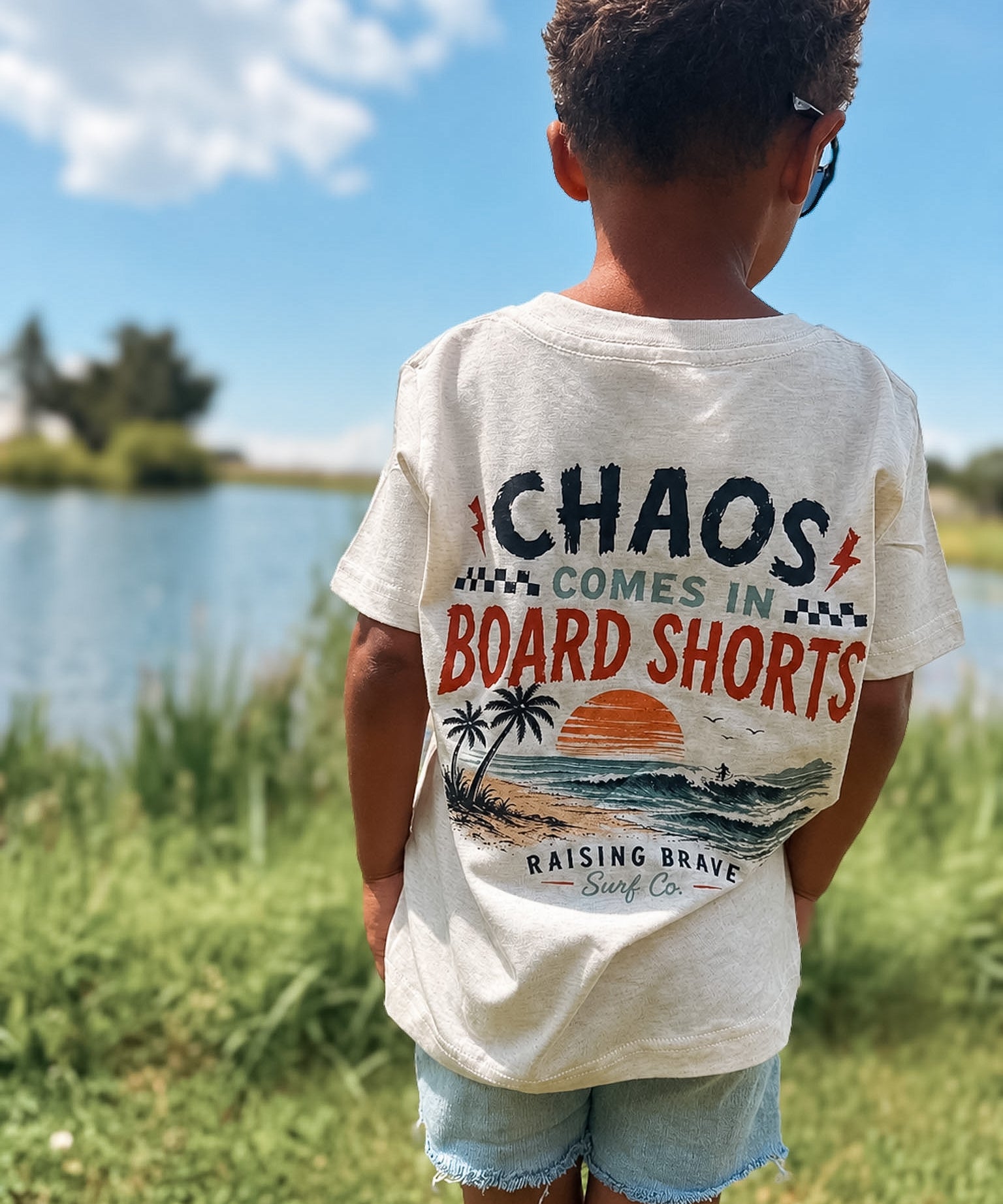 Child wearing a t-shirt with a graphic design and text, standing outdoors near a body of water.