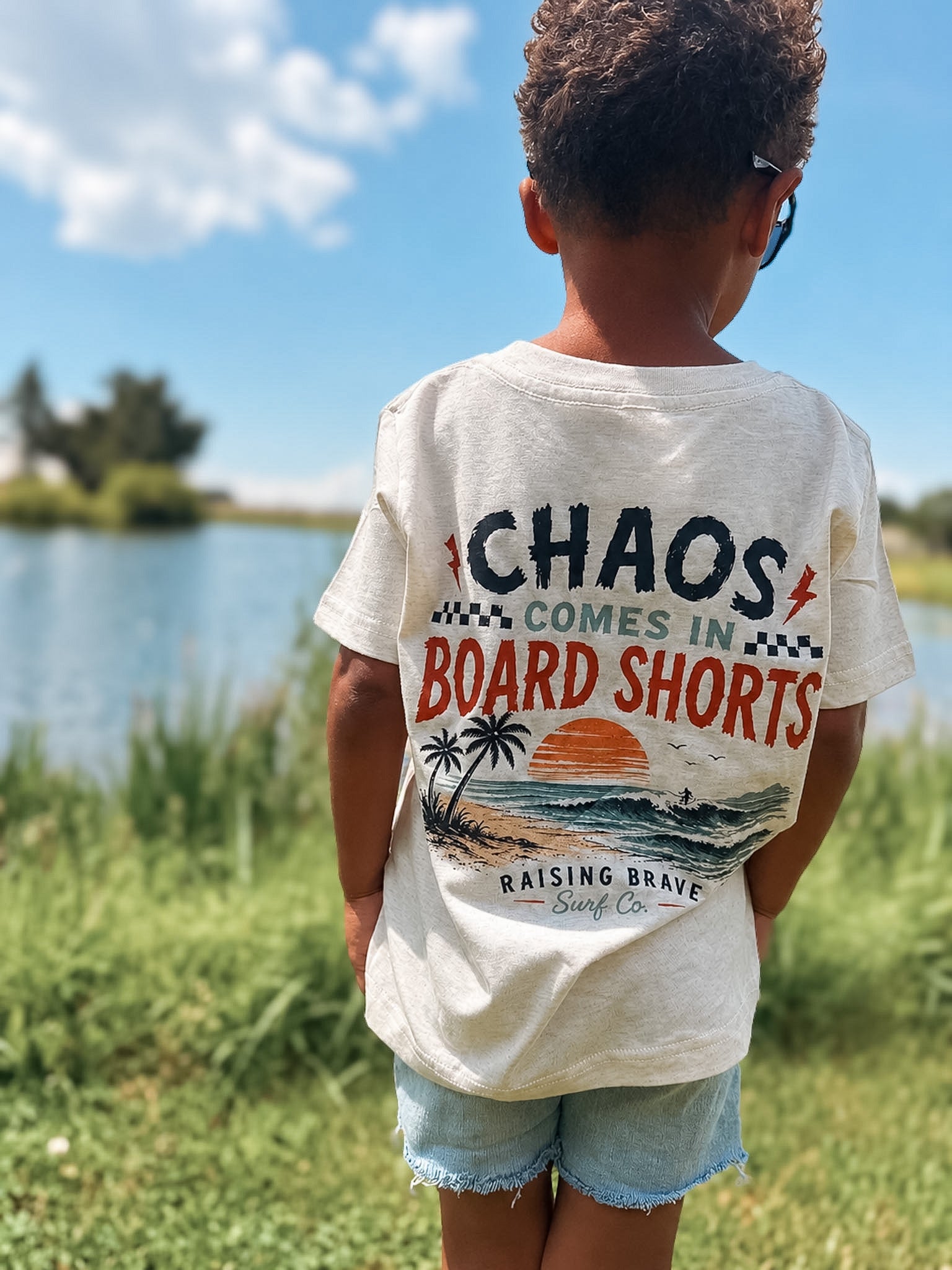 Child wearing a t-shirt with a graphic design and text, standing outdoors near a body of water.