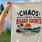 T-shirt with 'Chaos Comes in Board Shorts' design held by a person outdoors.