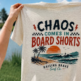 T-shirt with 'Chaos Comes in Board Shorts' design held by a person outdoors.