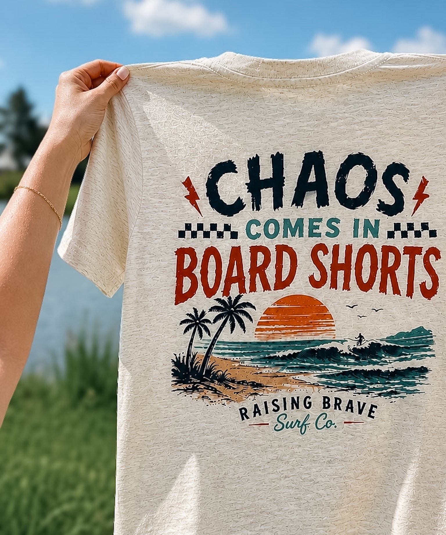 T-shirt with 'Chaos Comes in Board Shorts' design held by a person outdoors.