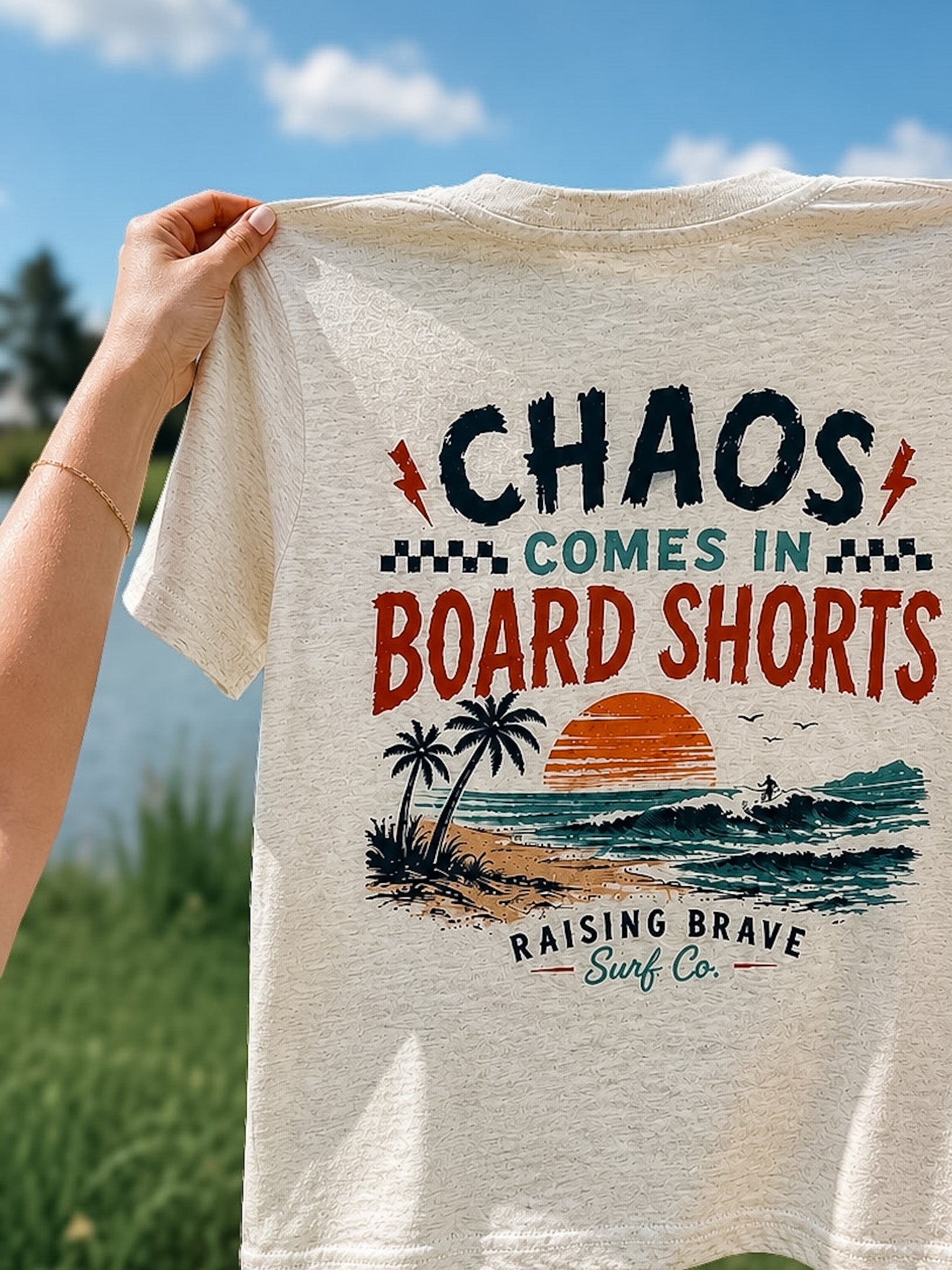 T-shirt with 'Chaos Comes in Board Shorts' design held by a person outdoors.