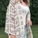 Woman wearing a patterned beige and white lace cardigan over a white top and jeans outdoors.