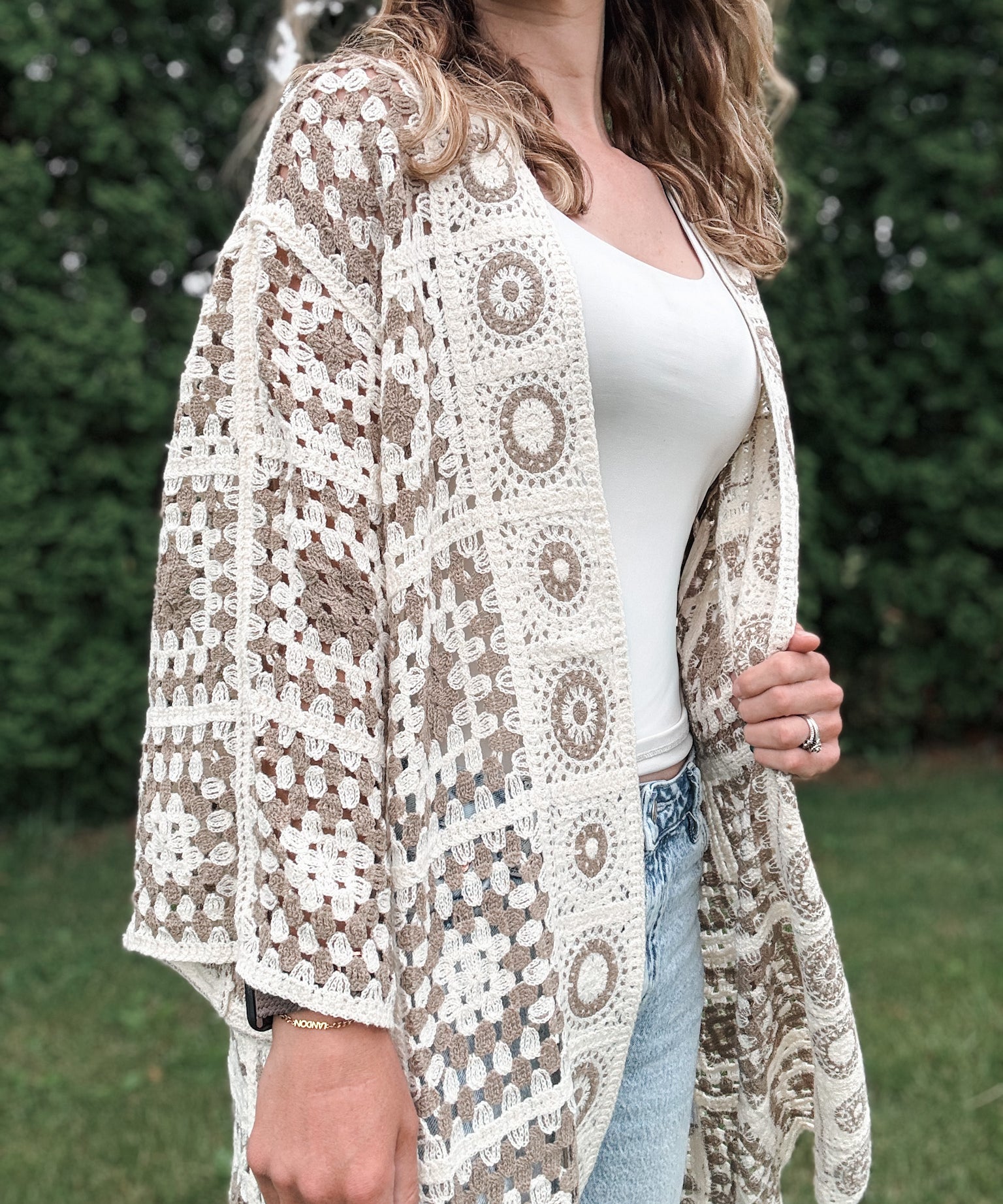 Woman wearing a patterned beige and white lace cardigan over a white top and jeans outdoors.