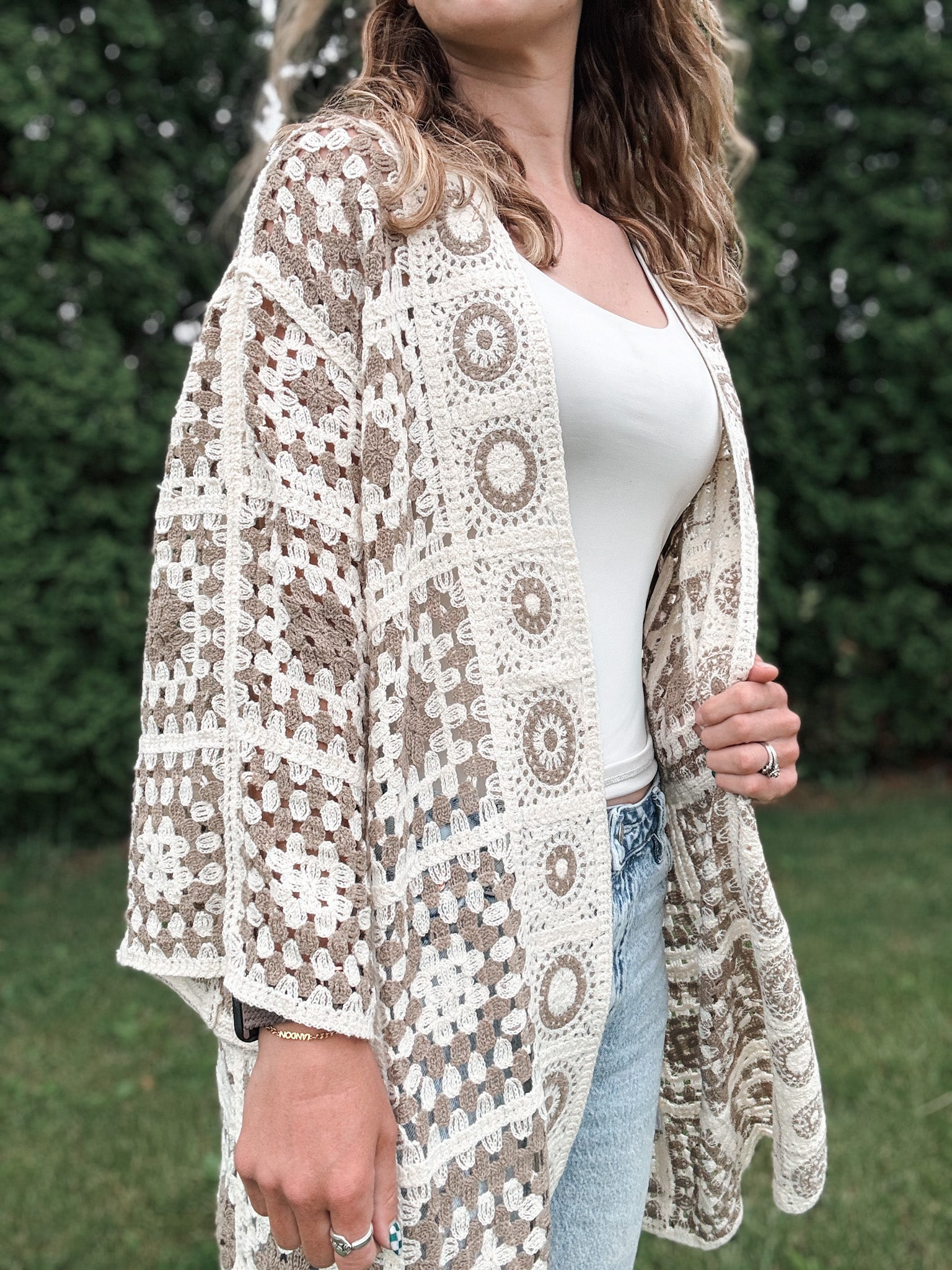 Woman wearing a patterned beige and white lace cardigan over a white top and jeans outdoors.