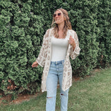 Woman wearing a white lace cardigan, white top, and light blue jeans standing in front of green bushes.