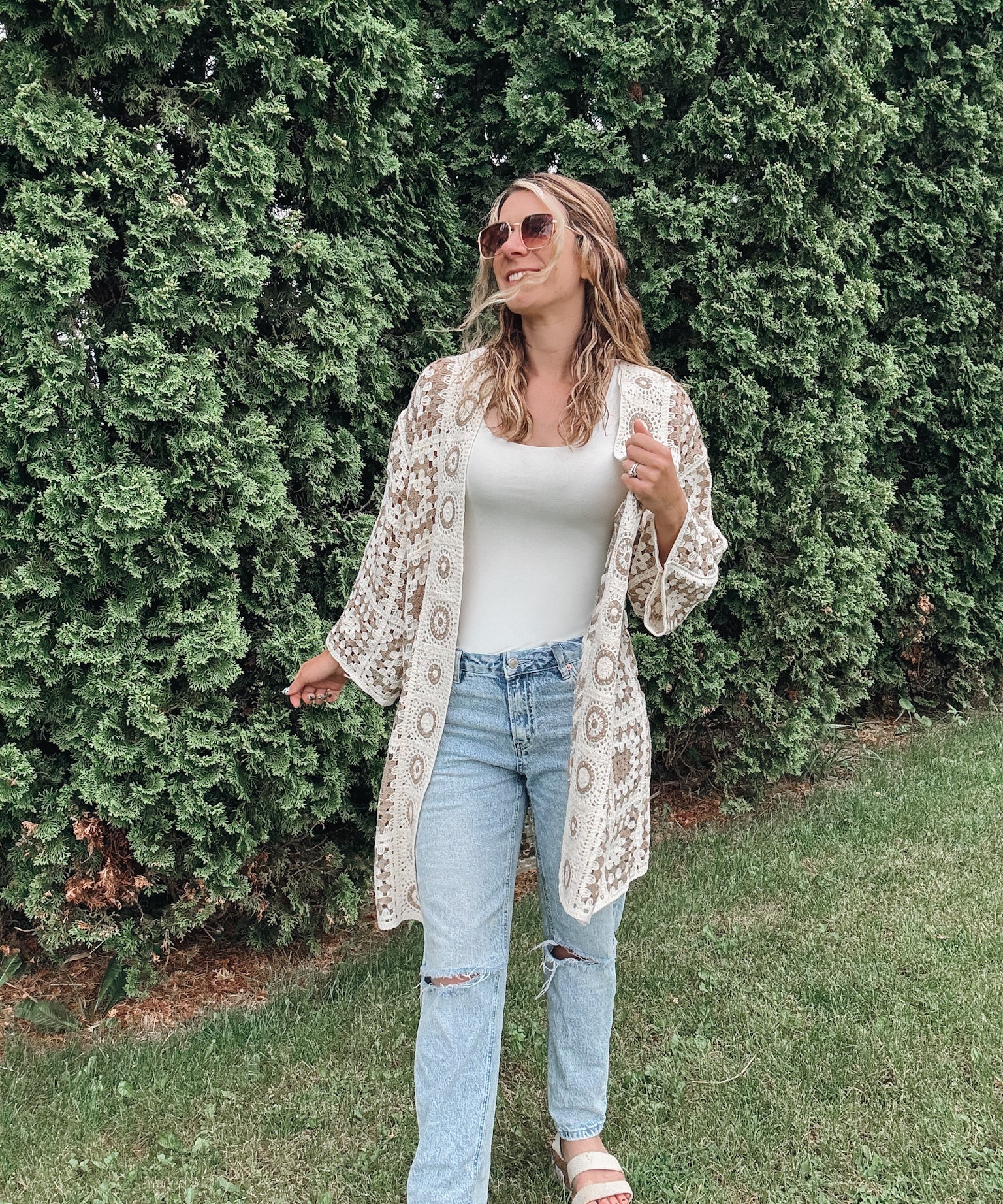 Woman wearing a white lace cardigan, white top, and light blue jeans standing in front of green bushes.