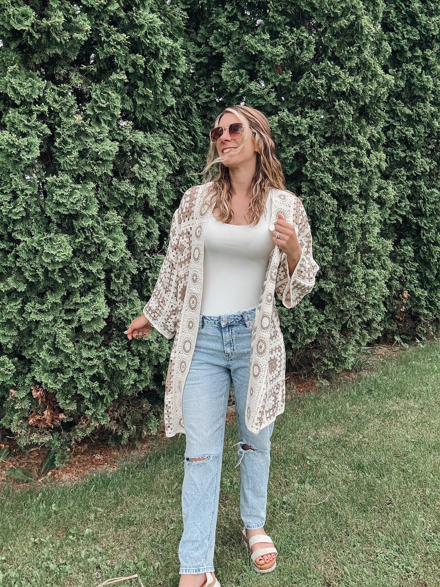 Woman wearing a white lace cardigan, white top, and light blue jeans standing in front of green bushes.