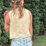 Person wearing a beige sleeveless top and light blue jeans standing against a green bush.