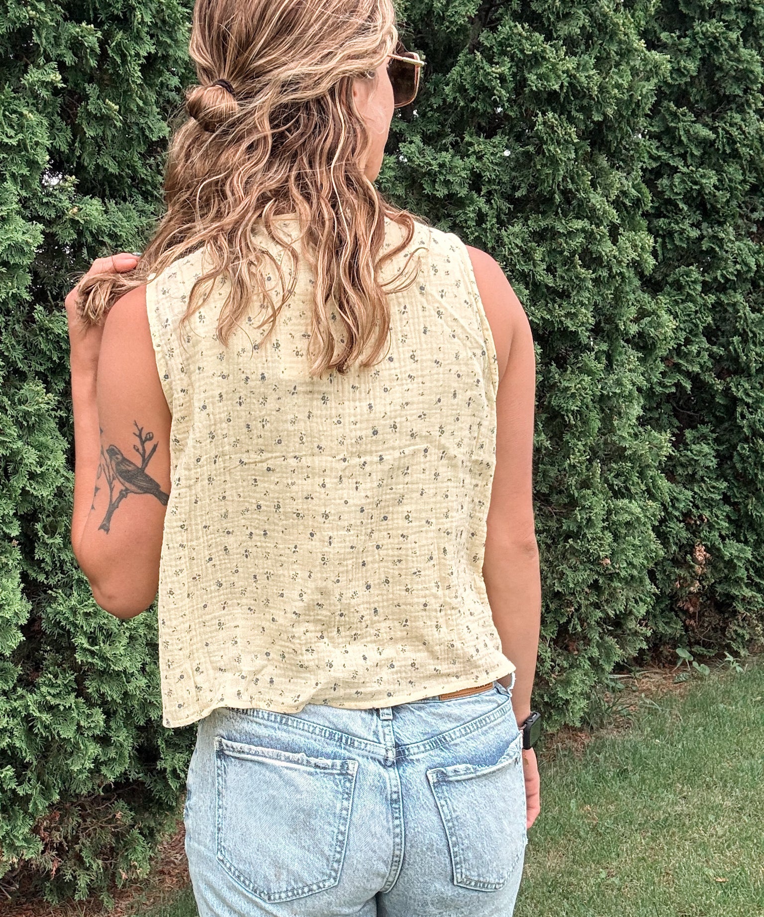 Person wearing a beige sleeveless top and light blue jeans standing against a green bush.