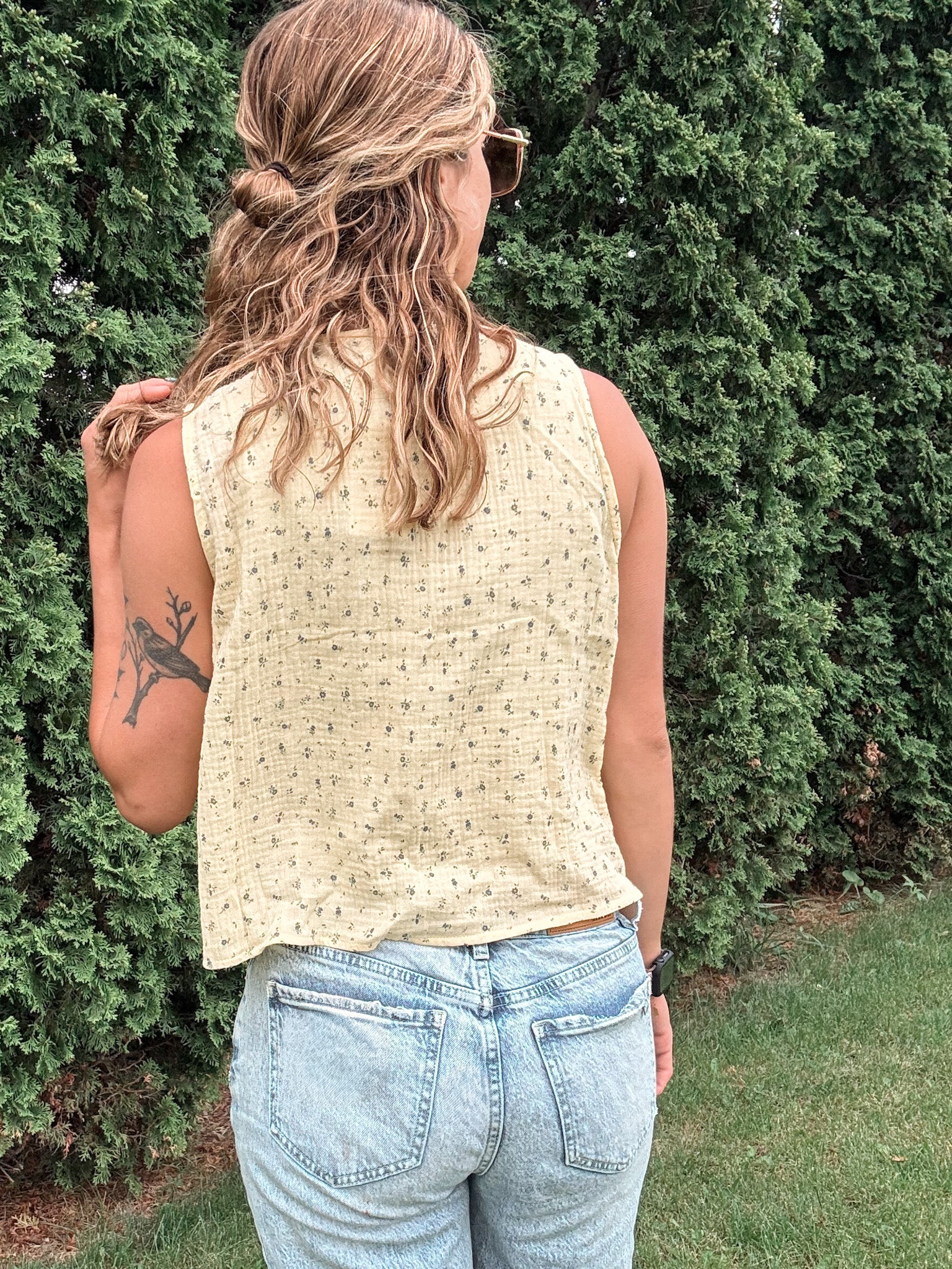 Person wearing a beige sleeveless top and light blue jeans standing against a green bush.