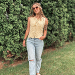 Woman wearing a sleeveless top and jeans standing in front of green bushes.