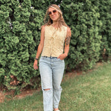 Woman wearing a sleeveless top and jeans standing in front of green bushes.