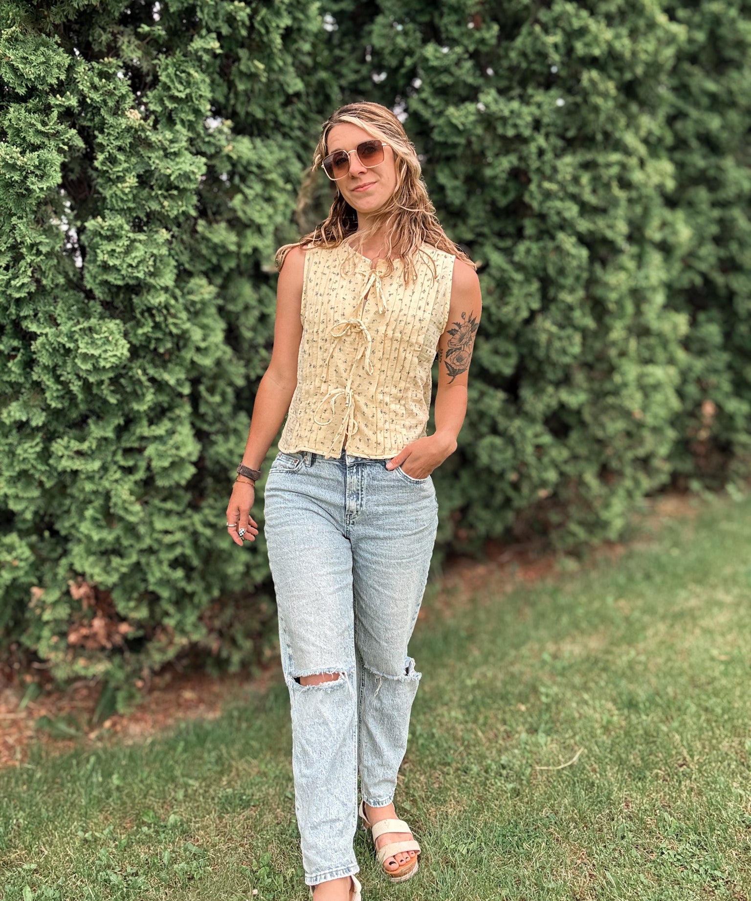 Woman wearing a sleeveless top and jeans standing in front of green bushes.