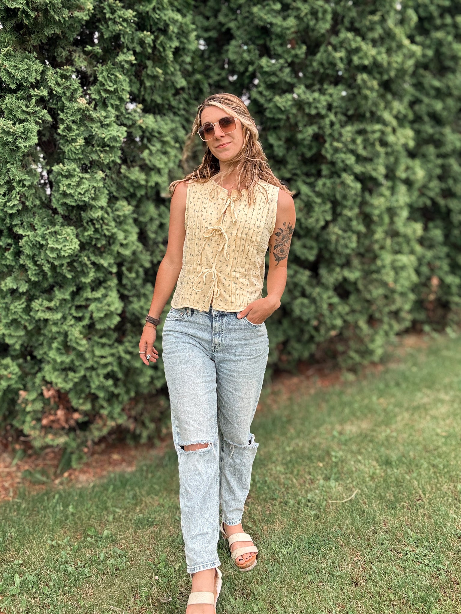Woman wearing a sleeveless top and jeans standing in front of green bushes.