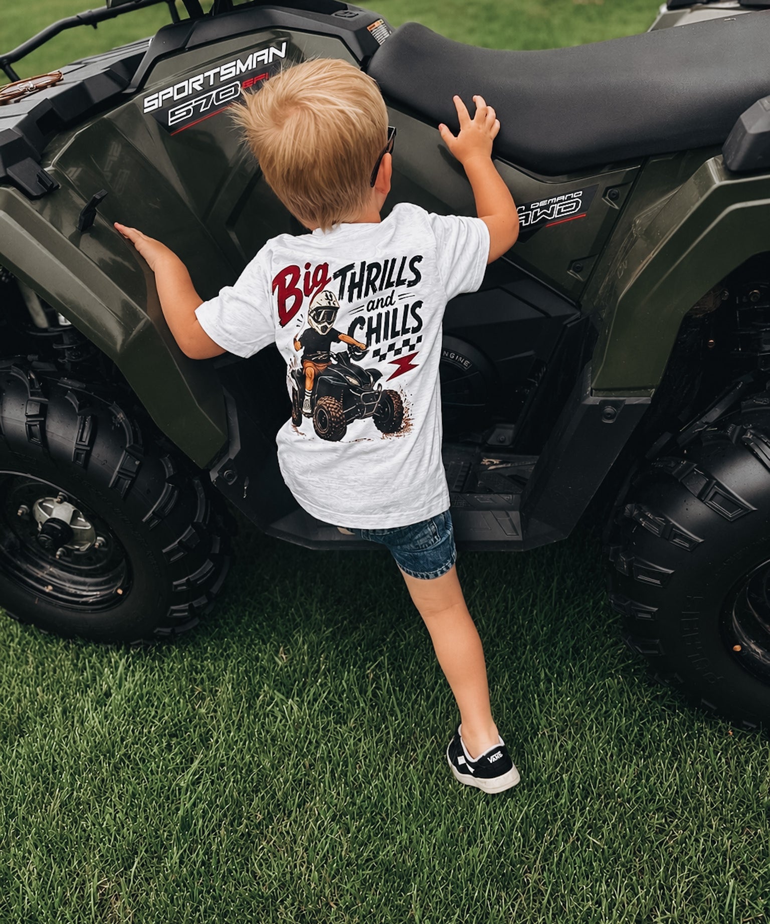 Child wearing a t-shirt with an ATV graphic standing next to a four-wheeler on grass