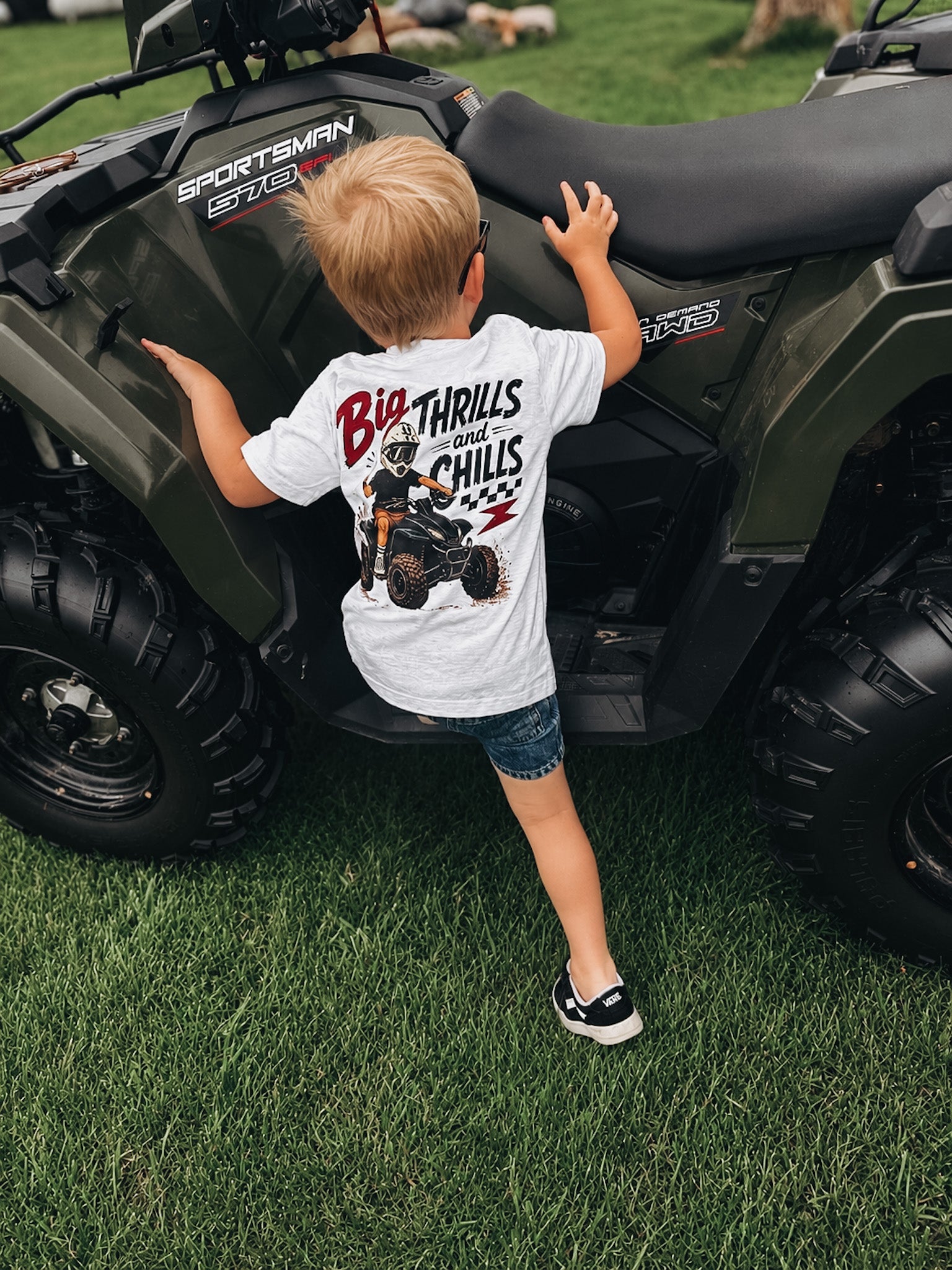 Child wearing a t-shirt with an ATV graphic standing next to a four-wheeler on grass