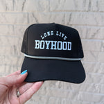 Black cap with 'Long Live Boyhood' text held against a light brick wall.