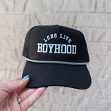 Black cap with 'Long Live Boyhood' text held against a light brick wall.