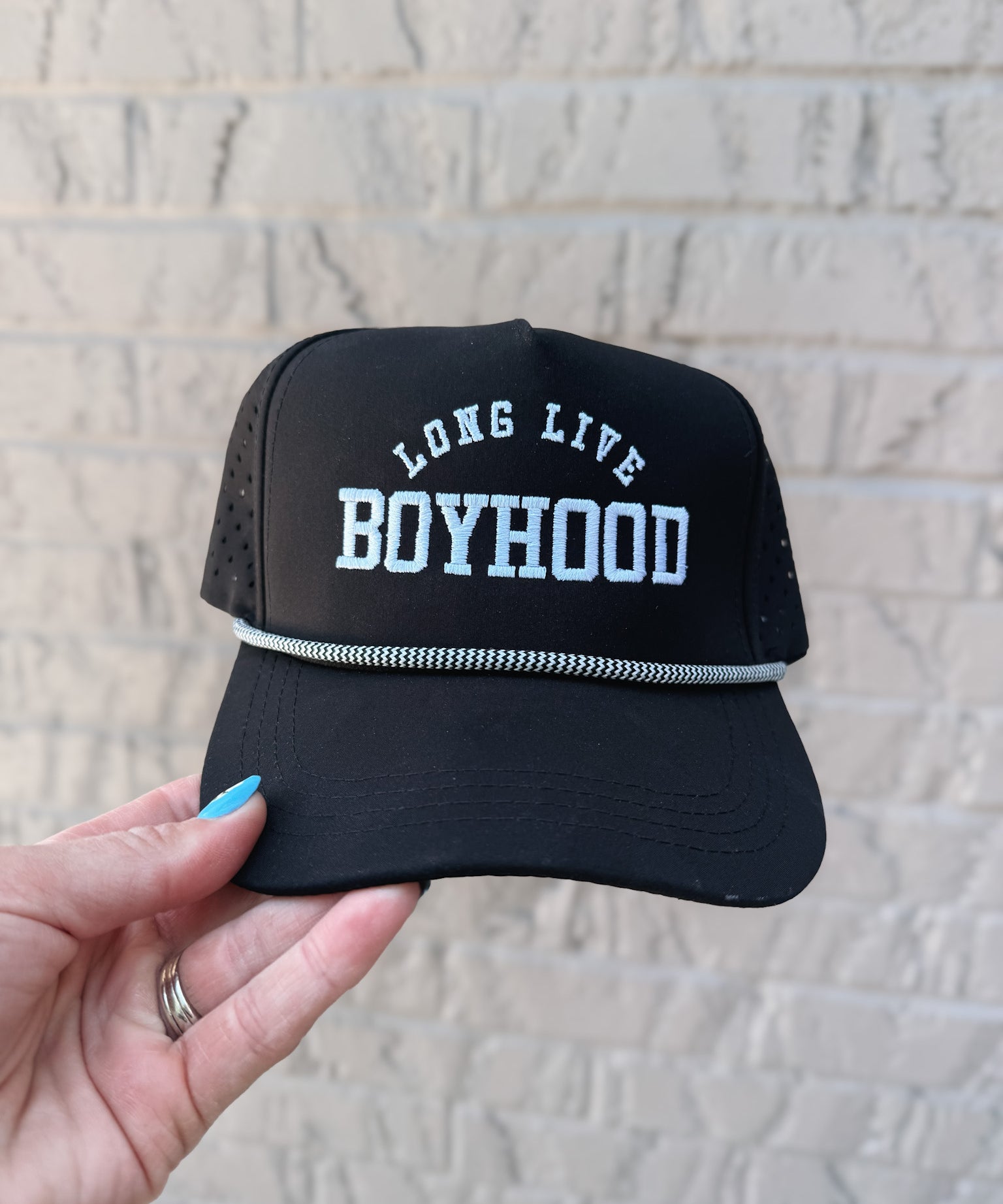 Black cap with 'Long Live Boyhood' text held against a light brick wall.