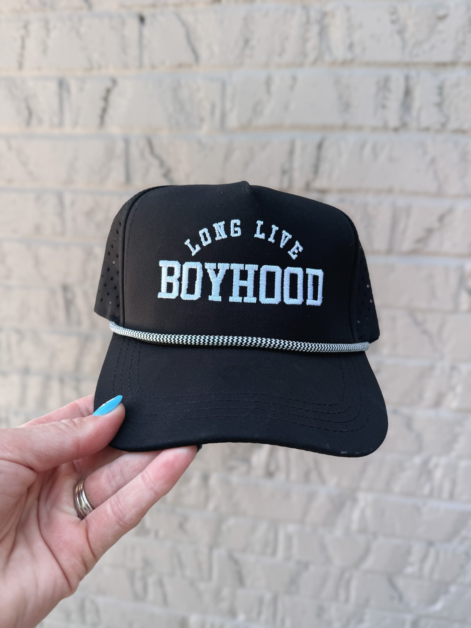 Black cap with 'Long Live Boyhood' text held against a light brick wall.