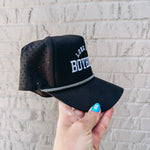 Black cap with mesh back held by a hand against a light gray brick wall.
