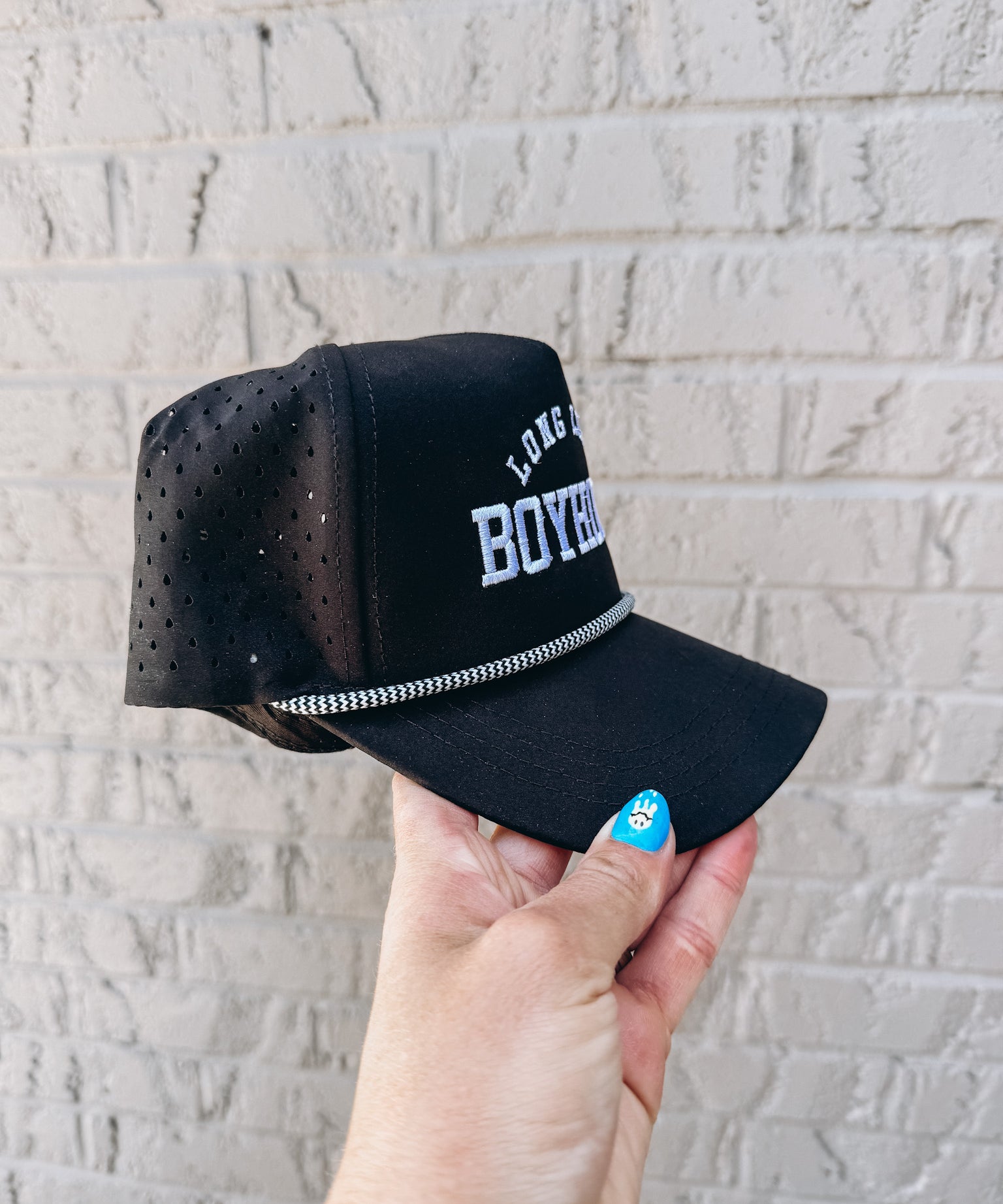 Black cap with mesh back held by a hand against a light gray brick wall.