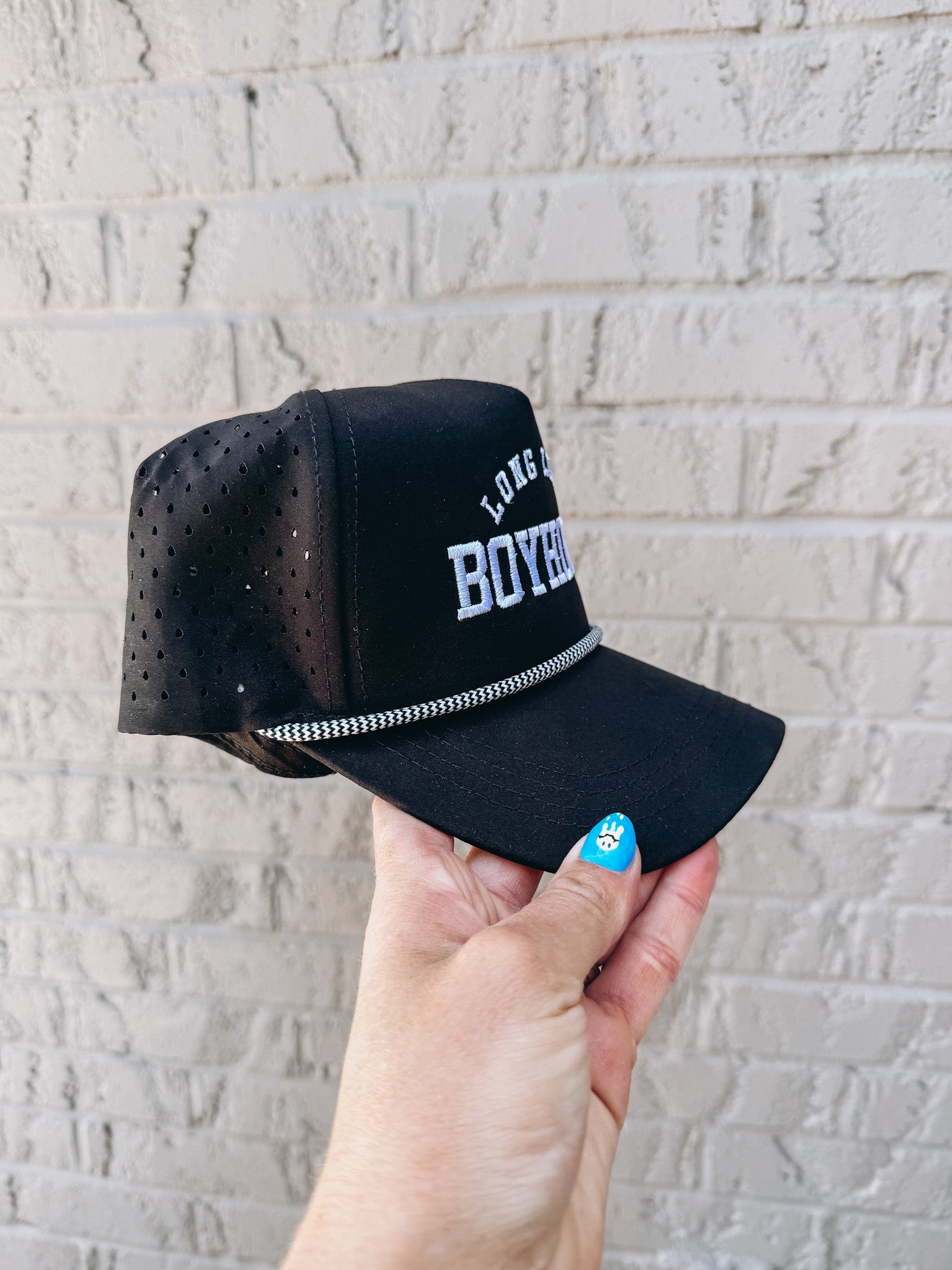 Black cap with mesh back held by a hand against a light gray brick wall.