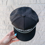 Black cap with perforated design held by a hand against a light gray brick wall.