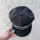 Black cap with perforated design held by a hand against a light gray brick wall.