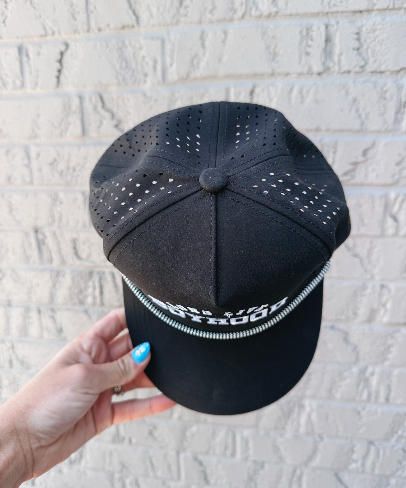 Black cap with perforated design held by a hand against a light gray brick wall.
