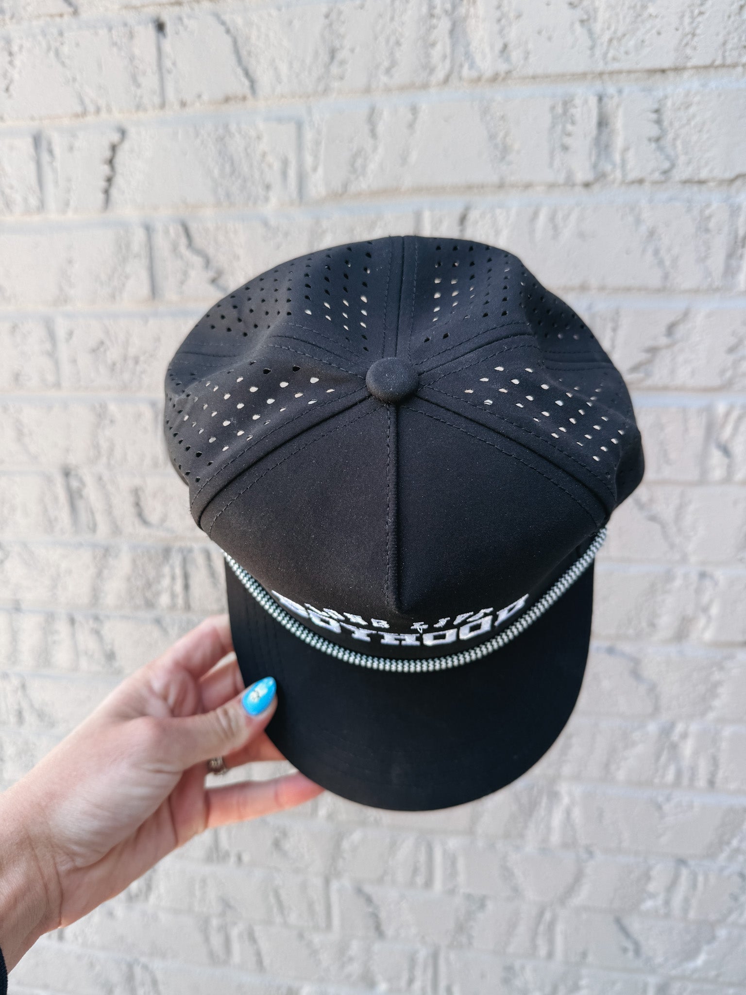 Black cap with perforated design held by a hand against a light gray brick wall.