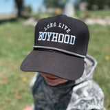 Child wearing a black cap with 'Long Live Boyhood' text in a park setting