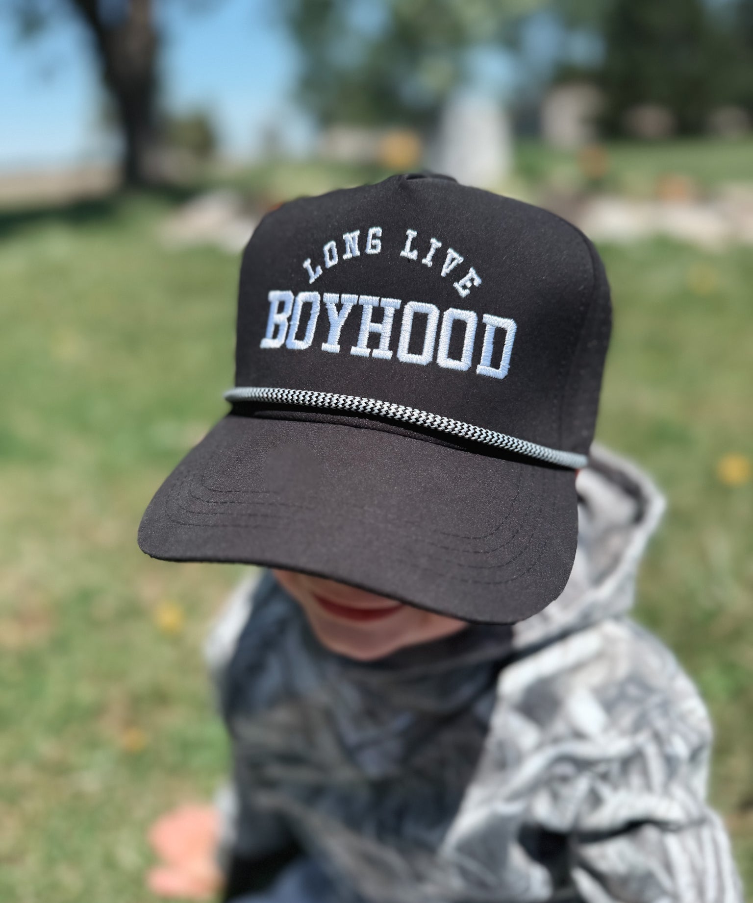 Child wearing a black cap with 'Long Live Boyhood' text in a park setting